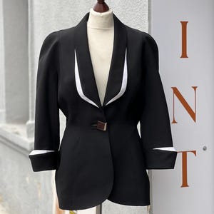 May include: A black blazer with white trim on the lapels and cuffs, displayed on a mannequin. The blazer has a single button closure and three-quarter sleeves. The background includes a sign with the word "VINTAGE".