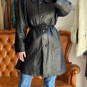 May include: A black leather coat with a belted waist, button closures, and a collared neckline. The coat has long sleeves and is paired with blue jeans. The coat is a vintage style.