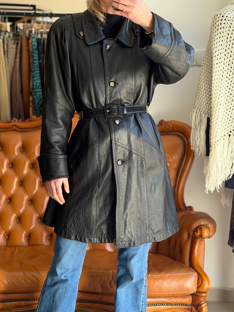 May include: A black leather coat with a belted waist, button closures, and a collared neckline. The coat has long sleeves and is paired with blue jeans. The coat is a vintage style.