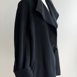 Pu&ograve; includere: Un cappotto nero in lana con ampio colletto e polsini risvoltati. Il cappotto presenta un design unico con un davanti drappeggiato e un sottile dettaglio a nodo sul lato. Il cappotto &egrave; esposto su un manichino.