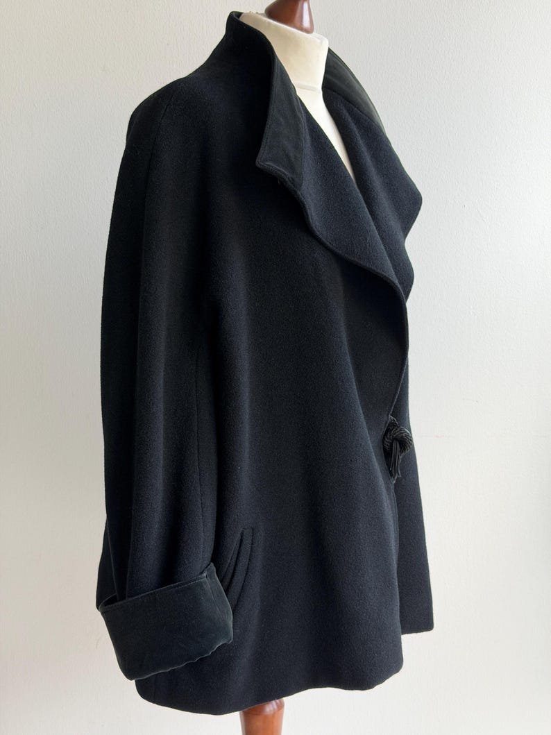 Pu&ograve; includere: Un cappotto nero in lana con ampio colletto e polsini risvoltati. Il cappotto presenta un design unico con un davanti drappeggiato e un sottile dettaglio a nodo sul lato. Il cappotto &egrave; esposto su un manichino.