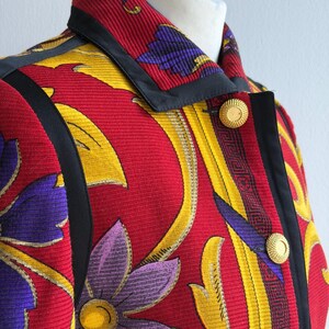 May include: A vibrant, patterned jacket featuring a red background with yellow and purple floral designs. The jacket has a black trim, gold buttons, and a structured collar. The fabric has a textured appearance.