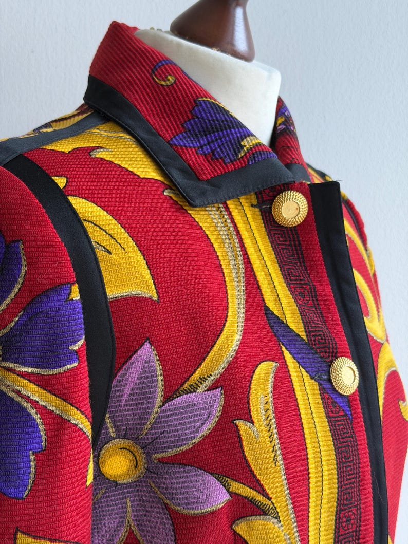 May include: A vibrant, patterned jacket featuring a red background with yellow and purple floral designs. The jacket has a black trim, gold buttons, and a structured collar. The fabric has a textured appearance.