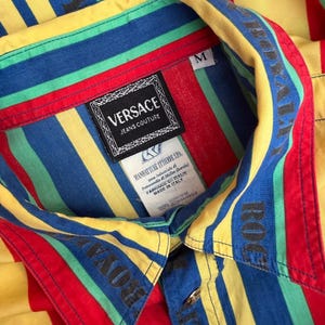 May include: A medium-sized, button-down shirt with bold, colorful stripes in red, yellow, blue, and green. The shirt features the "VERSACE JEANS COUTURE" label and the word "ROYAL" printed on the collar. The shirt is made of 100% cotton.