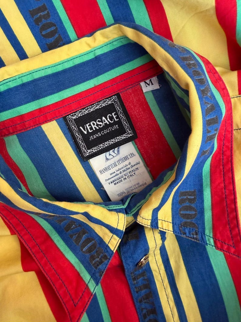 May include: A medium-sized, button-down shirt with bold, colorful stripes in red, yellow, blue, and green. The shirt features the "VERSACE JEANS COUTURE" label and the word "ROYAL" printed on the collar. The shirt is made of 100% cotton.