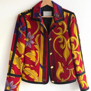 May include: A red, yellow, and black patterned jacket with floral and scroll designs. The jacket has a black collar and trim, gold buttons, and the brand name "VERSUS" on a label.