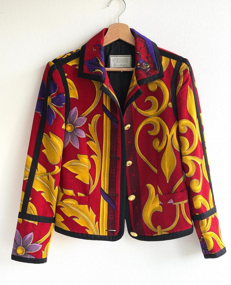 May include: A red, yellow, and black patterned jacket with floral and scroll designs. The jacket has a black collar and trim, gold buttons, and the brand name "VERSUS" on a label.