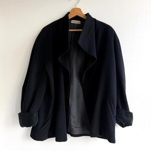 Pu&ograve; includere: Un cappotto nero in lana con ampio colletto e maniche con risvolto &egrave; appeso a una gruccia di legno. Il cappotto ha un design frontale aperto e fluido e una fodera scura. Il cappotto &egrave; esposto contro un muro bianco.