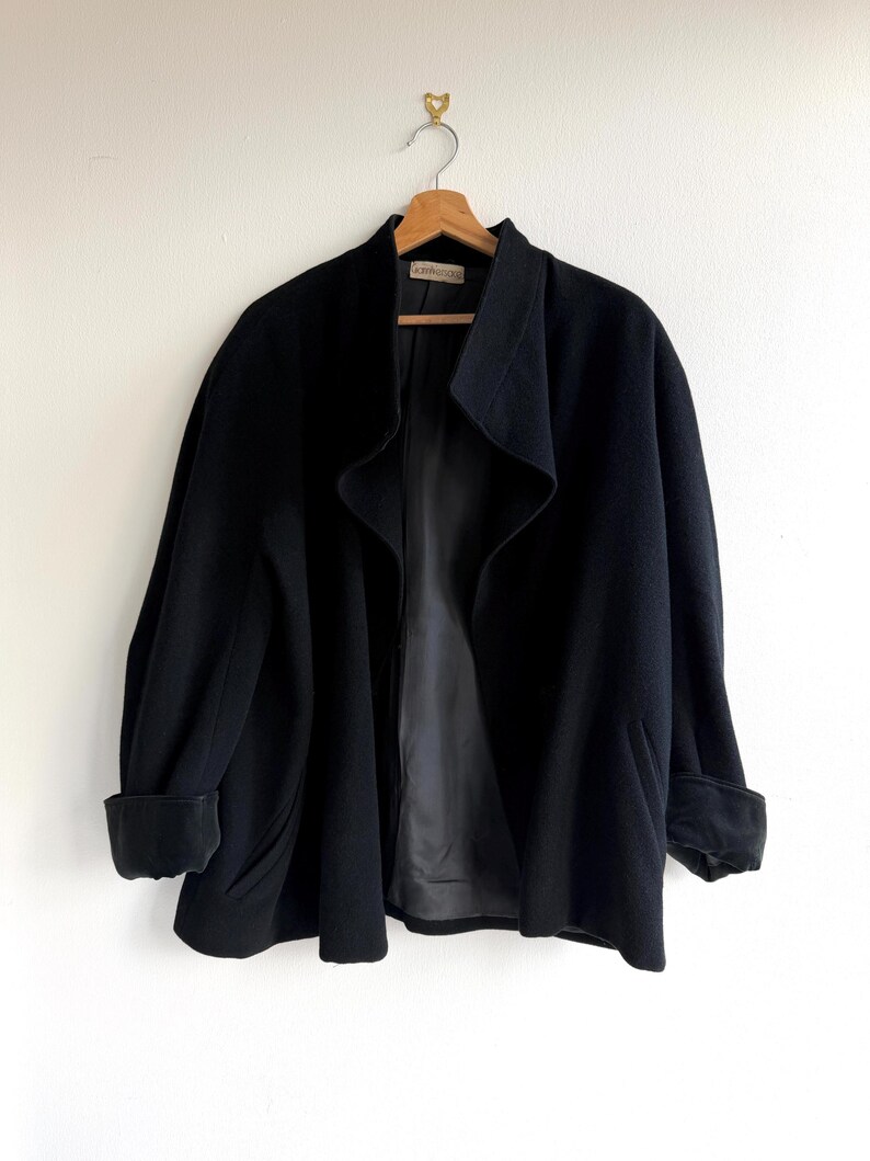 Pu&ograve; includere: Un cappotto nero in lana con ampio colletto e maniche con risvolto &egrave; appeso a una gruccia di legno. Il cappotto ha un design frontale aperto e fluido e una fodera scura. Il cappotto &egrave; esposto contro un muro bianco.