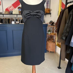May include: A black sleeveless dress with a satin bow detail at the waist. The dress is displayed on a wooden dress form. The background includes a blue wall, clothing racks, and decorative items.