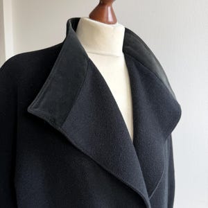 Pu&ograve; includere: Un cappotto nero con ampio colletto e revers &egrave; esposto su un manichino. Il colletto del cappotto ha una consistenza a contrasto e l'indumento &egrave; realizzato in un tessuto scuro e morbido. Il cappotto &egrave; uno stile classico.