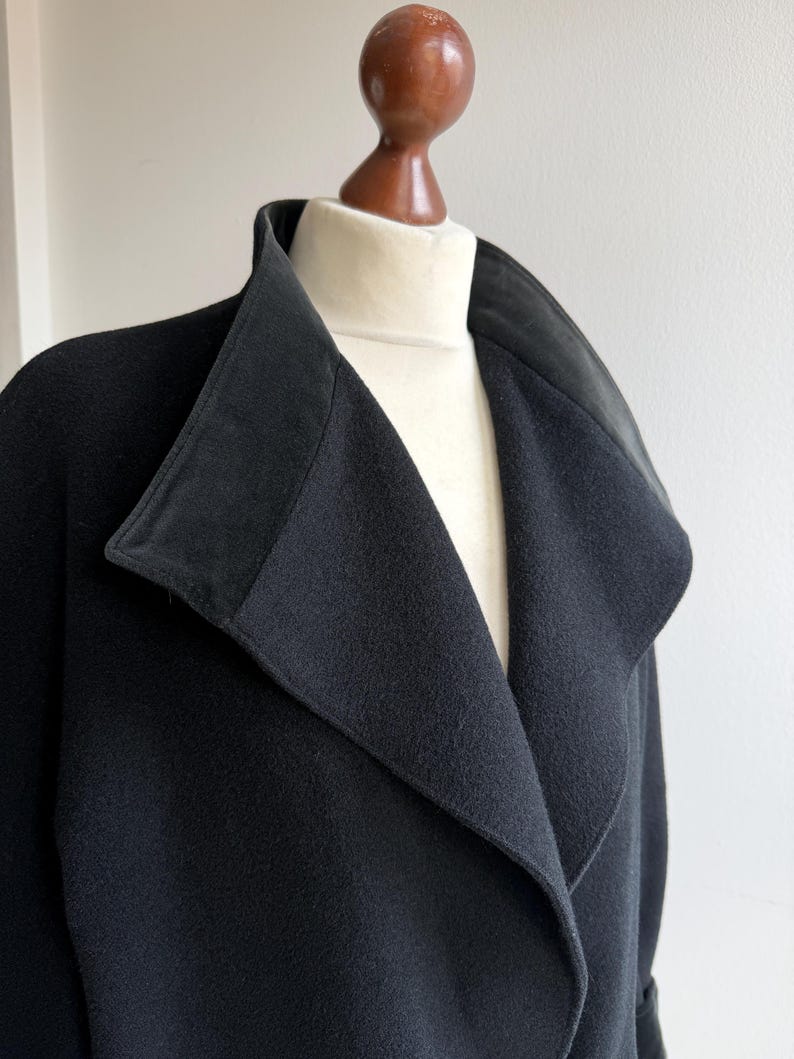 Pu&ograve; includere: Un cappotto nero con ampio colletto e revers &egrave; esposto su un manichino. Il colletto del cappotto ha una consistenza a contrasto e l'indumento &egrave; realizzato in un tessuto scuro e morbido. Il cappotto &egrave; uno stile classico.