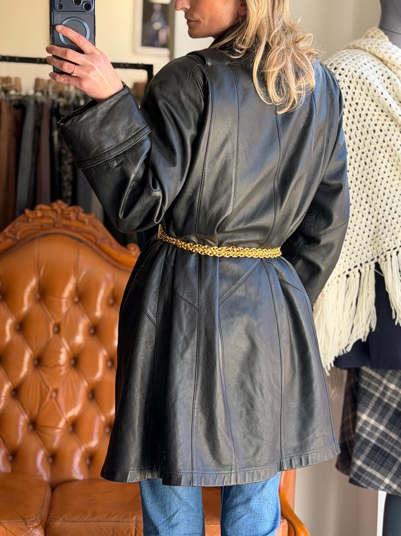 May include: A black leather coat with a gold chain belt. The coat has a flared skirt and long sleeves. The person is wearing blue jeans. The coat is a vintage style.