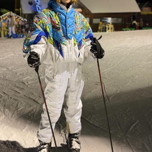 May include: A person wearing a white ski suit with a blue and colorful patterned jacket, black gloves, and a black beanie, holding ski poles. The person is standing on skis on a snowy slope at night.