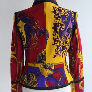 May include: A vibrant, patterned blazer with a striking color block design. The jacket features a combination of red, yellow, and purple hues, adorned with ornate floral and scroll motifs. Black trim outlines the collar, cuffs, and edges.