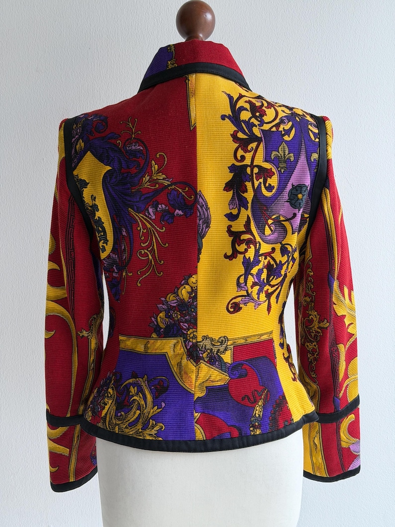 May include: A vibrant, patterned blazer with a striking color block design. The jacket features a combination of red, yellow, and purple hues, adorned with ornate floral and scroll motifs. Black trim outlines the collar, cuffs, and edges.