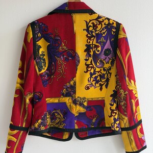 May include: A vintage blazer with a vibrant design. The jacket features a split design with red and yellow panels, adorned with ornate gold, purple, and black patterns. The blazer has a classic collar and black trim, hanging on a wooden hanger.