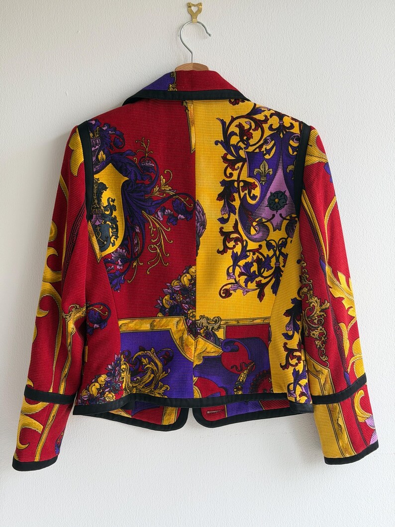 May include: A vintage blazer with a vibrant design. The jacket features a split design with red and yellow panels, adorned with ornate gold, purple, and black patterns. The blazer has a classic collar and black trim, hanging on a wooden hanger.
