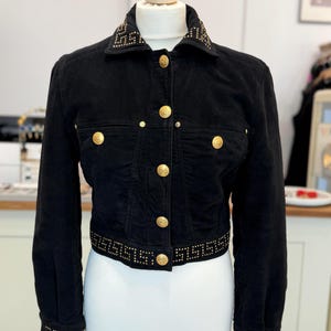 May include: A black cropped jacket featuring gold-tone metal buttons down the front and on the chest pockets. The collar, cuffs, and bottom hem are decorated with a gold stud pattern in a geometric Greek key design on the black fabric.