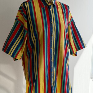 May include: A button-down shirt with vertical stripes in yellow, red, blue, and green. The short-sleeved shirt is displayed on a mannequin. The shirt has a classic collar and button closure.