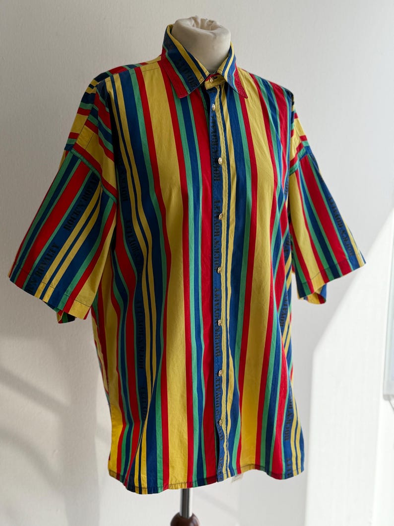 May include: A button-down shirt with vertical stripes in yellow, red, blue, and green. The short-sleeved shirt is displayed on a mannequin. The shirt has a classic collar and button closure.