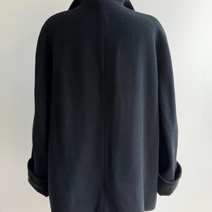 Pu&ograve; includere: Un cappotto nero in lana con ampio colletto e polsini risvoltati. Il cappotto &egrave; esposto su un manichino di legno su sfondo bianco. Il design del cappotto &egrave; semplice ed elegante, con una silhouette classica.