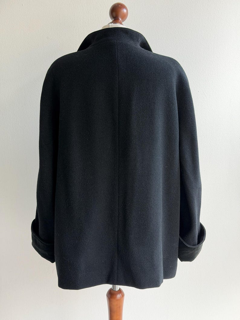 Pu&ograve; includere: Un cappotto nero in lana con ampio colletto e polsini risvoltati. Il cappotto &egrave; esposto su un manichino di legno su sfondo bianco. Il design del cappotto &egrave; semplice ed elegante, con una silhouette classica.