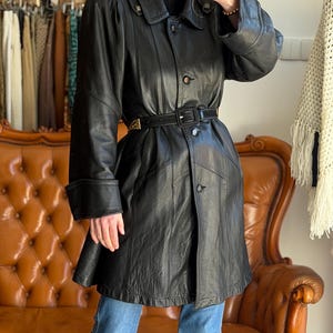 May include: A black leather coat with a belted waist and button closures. The coat has a classic design with a collar and long sleeves. The coat is worn over blue jeans. The coat is approximately 40 inches long.