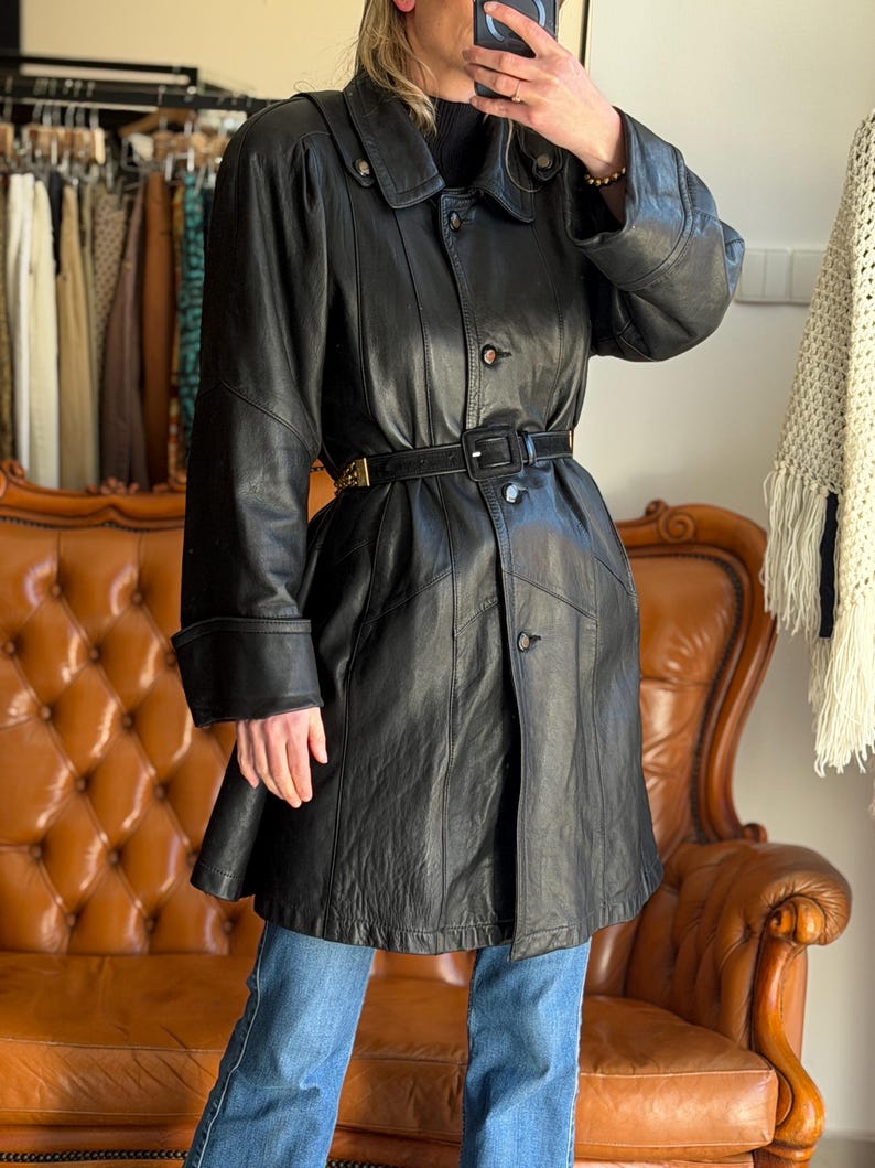 May include: A black leather coat with a belted waist and button closures. The coat has a classic design with a collar and long sleeves. The coat is worn over blue jeans. The coat is approximately 40 inches long.
