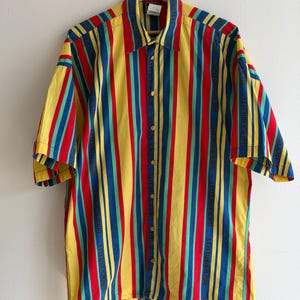 May include: A short-sleeved button-down shirt with vertical stripes in yellow, red, blue, and green. The shirt is hanging on a wooden hanger against a white wall. The shirt has a collar and gold-colored buttons.