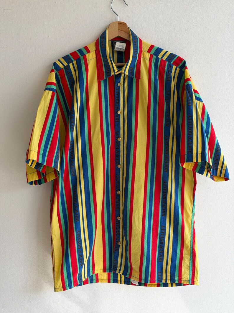 May include: A short-sleeved button-down shirt with vertical stripes in yellow, red, blue, and green. The shirt is hanging on a wooden hanger against a white wall. The shirt has a collar and gold-colored buttons.
