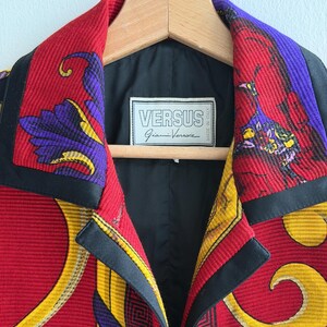 May include: A close-up of a Versus Gianni Versace jacket. The jacket features a vibrant red, yellow, and purple pattern with black trim. The label inside the jacket reads "VERSUS Gianni Versace MADE IN ITALY". The jacket is hanging on a wooden hanger.