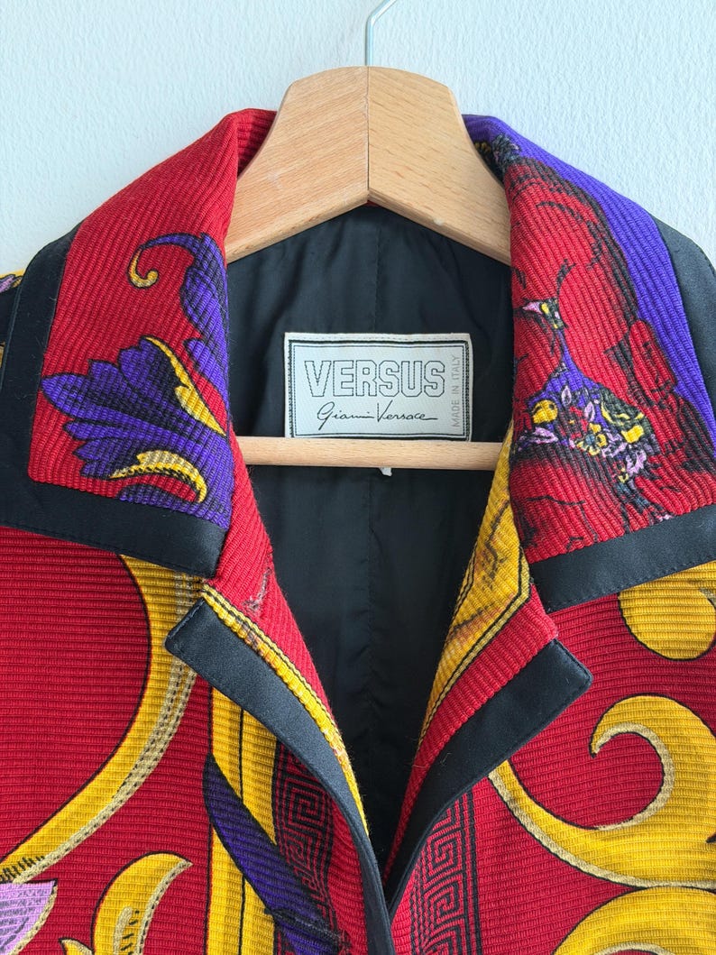 May include: A close-up of a Versus Gianni Versace jacket. The jacket features a vibrant red, yellow, and purple pattern with black trim. The label inside the jacket reads "VERSUS Gianni Versace MADE IN ITALY". The jacket is hanging on a wooden hanger.