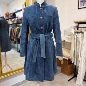 May include: A blue suede coat featuring a high collar, long sleeves, and a matching tie belt at the waist. The coat is secured with three round, metallic buttons down the front and includes vertical seam detailing along the bodice and skirt.