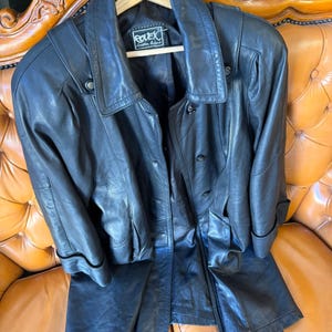 May include: A black leather jacket and matching pants set. The jacket has a collared neckline, button closures, and pockets. The pants are straight-legged. The set is displayed on a wooden hanger and a brown leather couch.