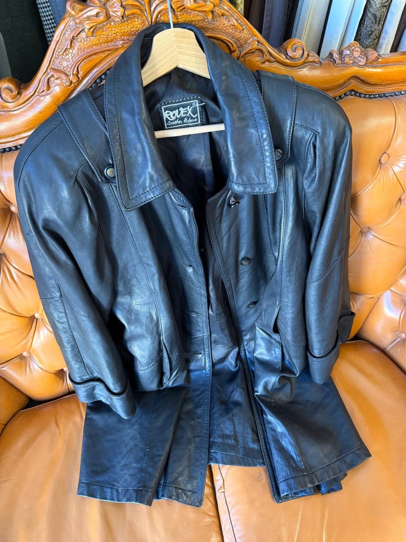 May include: A black leather jacket and matching pants set. The jacket has a collared neckline, button closures, and pockets. The pants are straight-legged. The set is displayed on a wooden hanger and a brown leather couch.