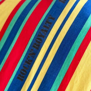 May include: Close-up of a fabric with bold, diagonal stripes in red, yellow, blue, and green. The words "ROCK'N ROYALTY" are printed in black along the stripes. The fabric appears to be part of a garment.