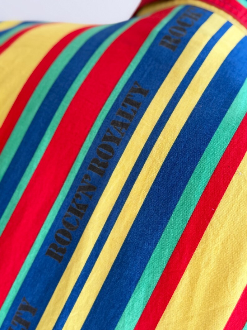 May include: Close-up of a fabric with bold, diagonal stripes in red, yellow, blue, and green. The words "ROCK'N ROYALTY" are printed in black along the stripes. The fabric appears to be part of a garment.