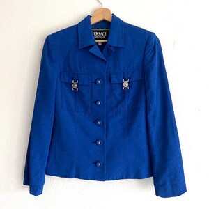 May include: A royal blue blazer with a button-up front and two flap pockets with silver hardware. The blazer has the text "VERSACE" and "COUTURE" on the label.