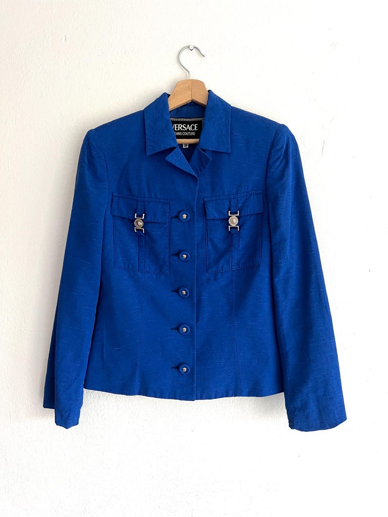 May include: A royal blue blazer with a button-up front and two flap pockets with silver hardware. The blazer has the text "VERSACE" and "COUTURE" on the label.