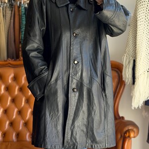 May include: A black leather coat with a button-up front, a collar, and pockets. The coat is knee-length and appears to be vintage. The coat is worn over a black sweater and blue jeans. The background includes a brown leather couch and a clothing rack.