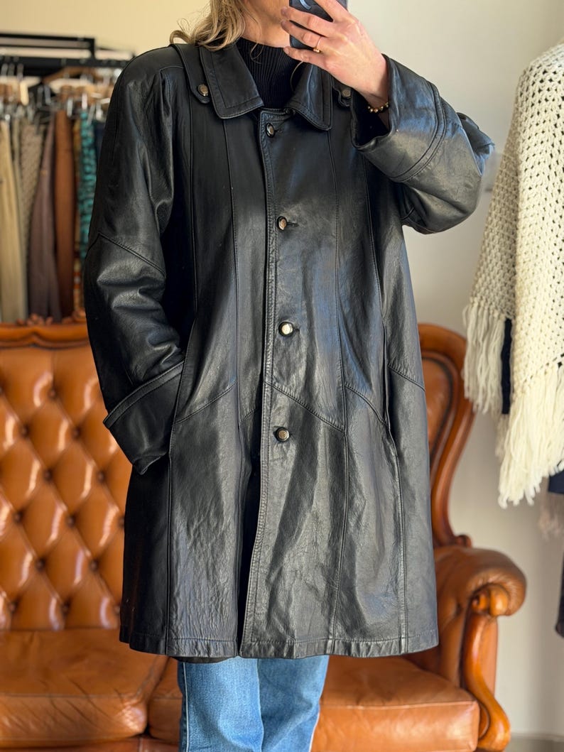 May include: A black leather coat with a button-up front, a collar, and pockets. The coat is knee-length and appears to be vintage. The coat is worn over a black sweater and blue jeans. The background includes a brown leather couch and a clothing rack.