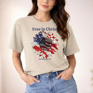 Può includere: T-shirt beige con un disegno di girasole con i colori della bandiera americana e la scritta "Free in Christ" sopra il fiore. Il testo "Galati 5:1" è sotto.