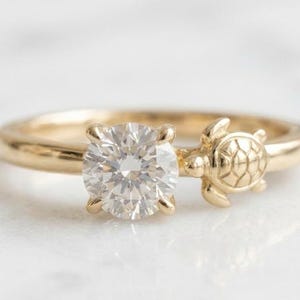 Può includere: Un anello in oro con una pietra preziosa rotonda, trasparente e sfaccettata in una montatura a quattro griffe, accanto a un piccolo ciondolo a forma di tartaruga marina in oro scolpito con un motivo a guscio dettagliato sulla fascia.