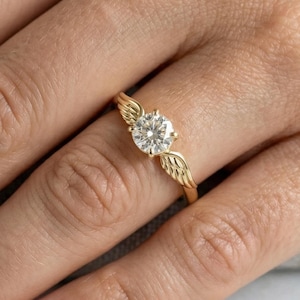 May include: A gold ring with a large, round, clear gemstone. The ring features a wing design on either side of the gemstone. The ring is on a person's finger, with a neutral background.