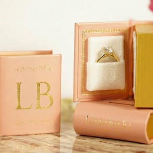 May include: A peach-colored ring box shaped like a book, with gold lettering and accents. The box is open, revealing a gold ring with a diamond. The initials "LB" are on the front of the closed box.