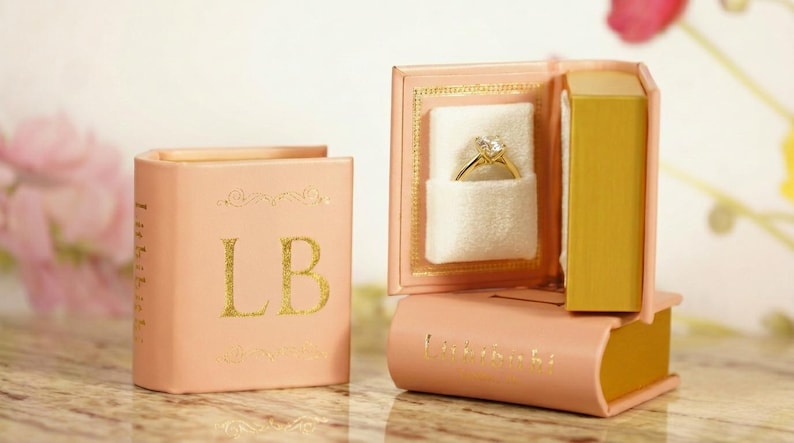 May include: A peach-colored ring box shaped like a book, with gold lettering and accents. The box is open, revealing a gold ring with a diamond. The initials "LB" are on the front of the closed box.