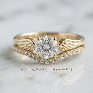 May include: A gold engagement ring with a large, round, clear gemstone and a matching wedding band. The engagement ring features wing-shaped details on the sides of the gemstone. The wedding band is curved and set with small, clear gemstones. Text: wedding band sold separately :)