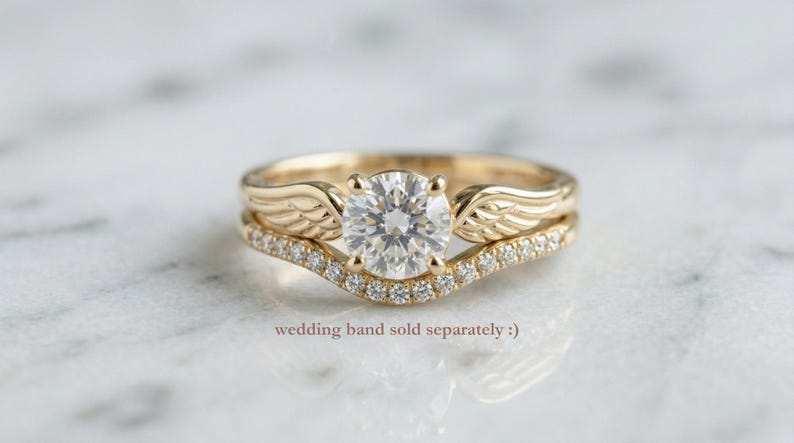 May include: A gold engagement ring with a large, round, clear gemstone and a matching wedding band. The engagement ring features wing-shaped details on the sides of the gemstone. The wedding band is curved and set with small, clear gemstones. Text: wedding band sold separately :)