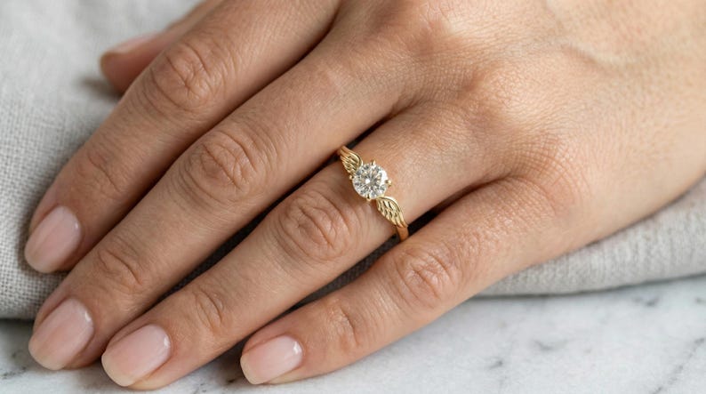 May include: A gold ring with a large, round, clear gemstone. The ring features a wing design on either side of the gemstone. The ring is on a person's finger, with a neutral background.